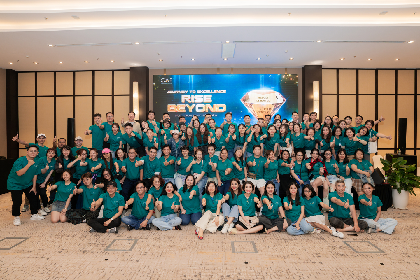 Tập thể nhân viên CAF mặc đồng phục xanh mòng két chụp ảnh kỷ niệm tại sự kiện Journey to Excellence - Rise Beyond