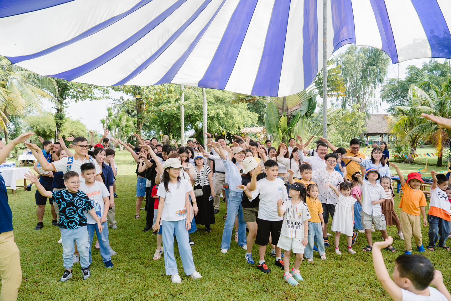 Nhóm người lớn và trẻ em tham gia hoạt động tập thể ngoài trời dưới mái che sọc xanh trắng