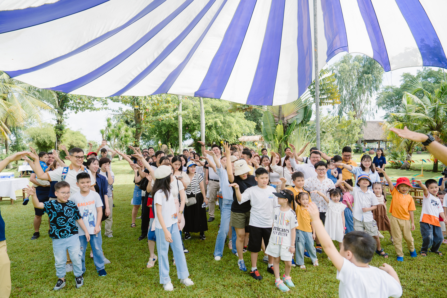 Nhóm người lớn và trẻ em tham gia hoạt động tập thể ngoài trời dưới mái che sọc xanh trắng